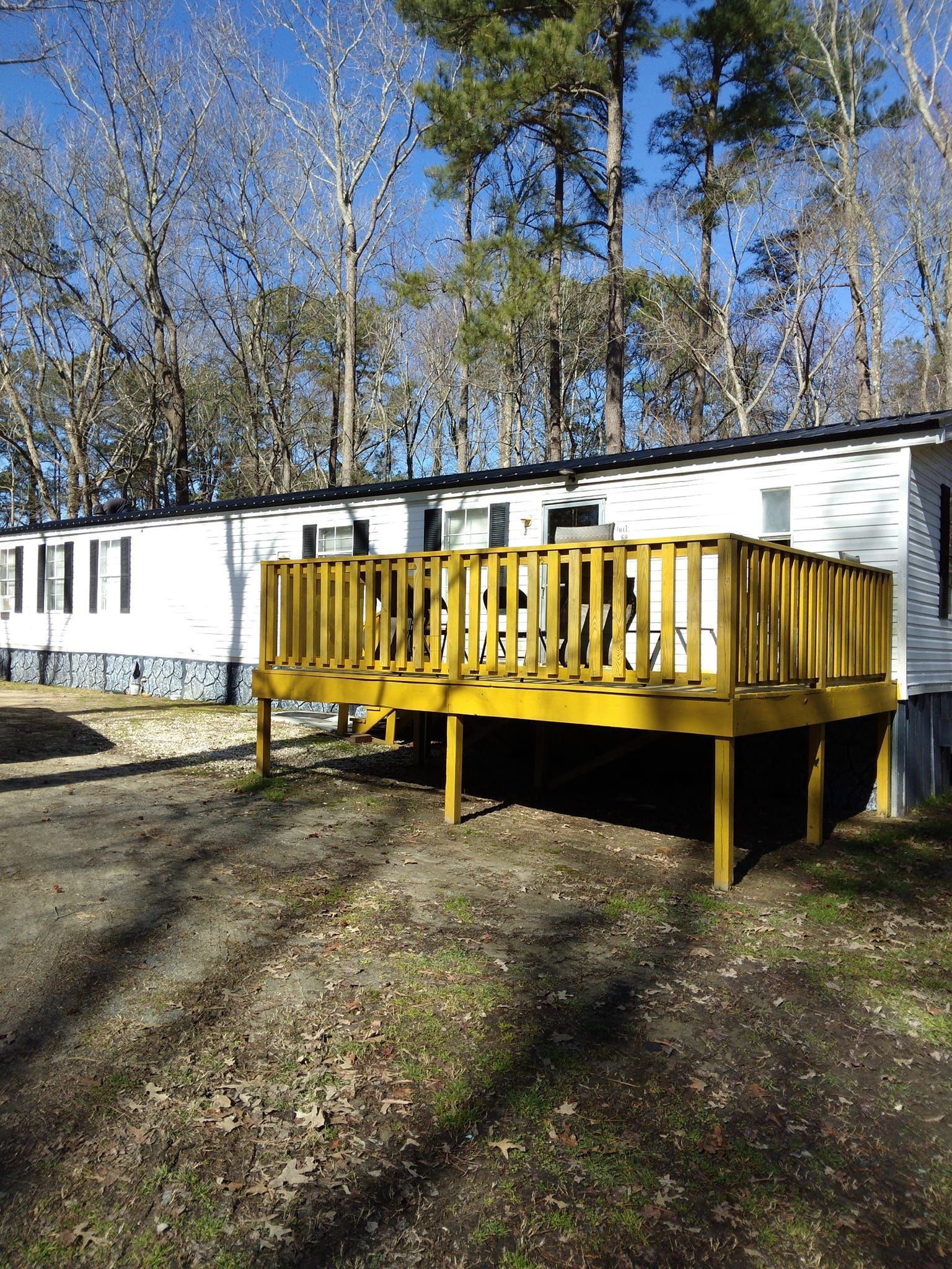 White mobile home with a bright yellow wooden deck in a wooded yard on a sunny day