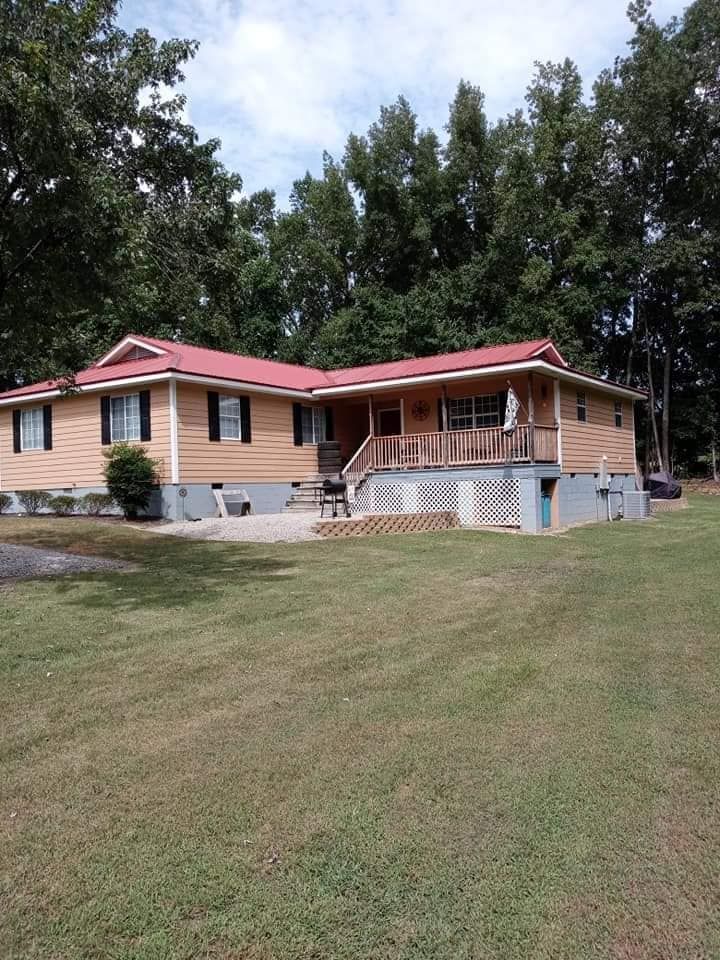 Single-story tan house with red roof and a covered porch on a grassy lawn, backed by trees.