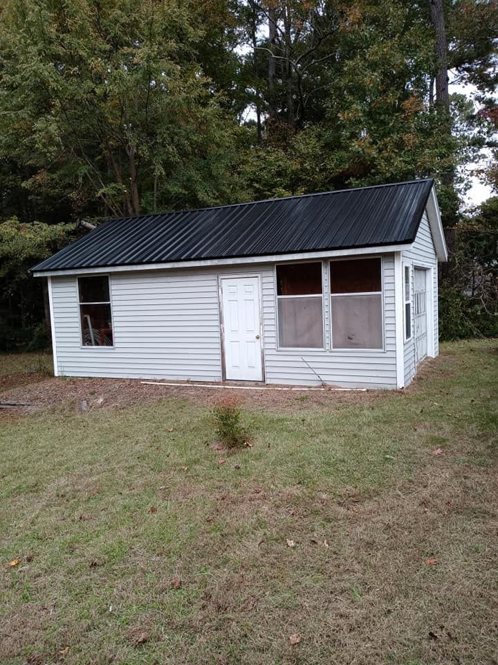 Small white shed with a black roof and boarded windows in a grassy yard near trees
