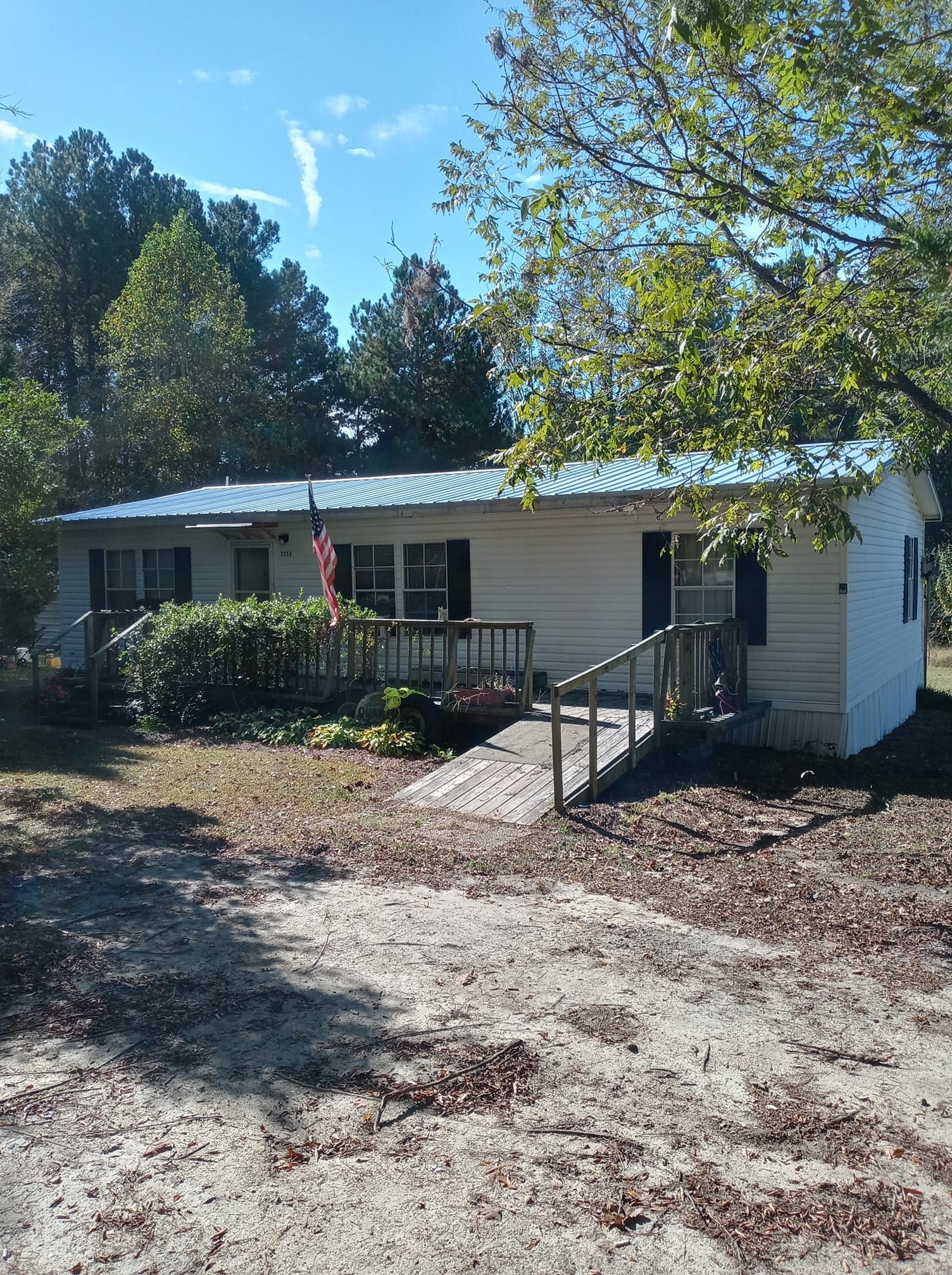 Single-story white house with porch, ramp, and trees in a sunny yard