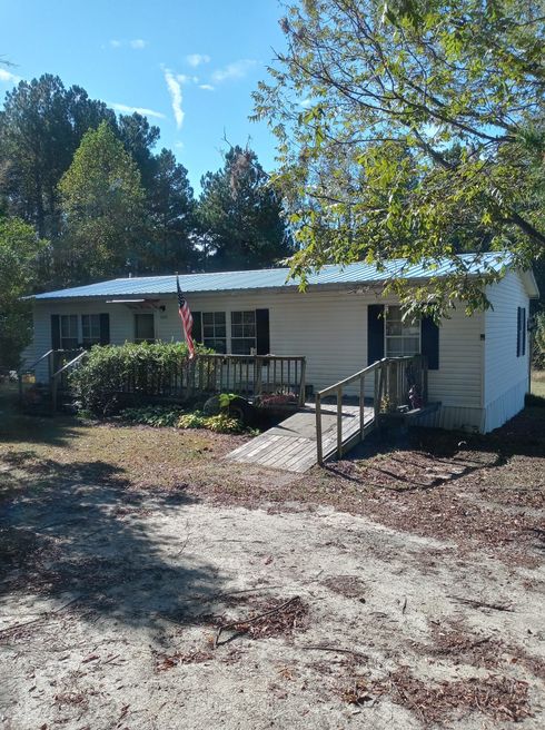 Single-story white house with porch, ramp, and trees in a sunny yard