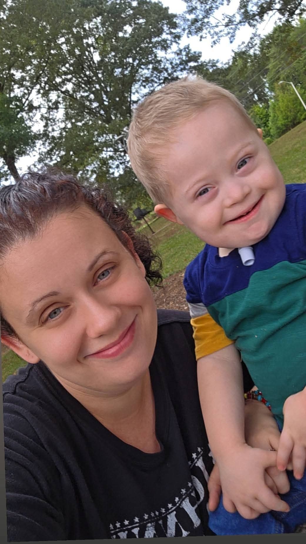 Two people smiling outdoors, one holding a child in a blue and green shirt.