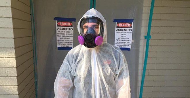 Person in white hazmat suit and respirator mask standing outside a building with warning signs.