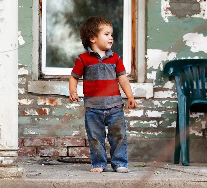 Small child in a red and gray shirt standing on a porch beside a weathered wall and chair