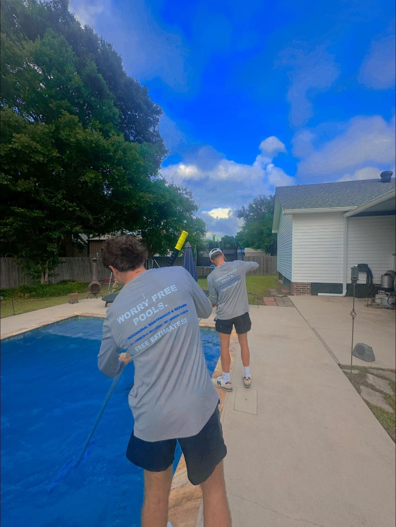 Two people in gray long-sleeved shirts clean a blue swimming pool in a sunny residential backyard.