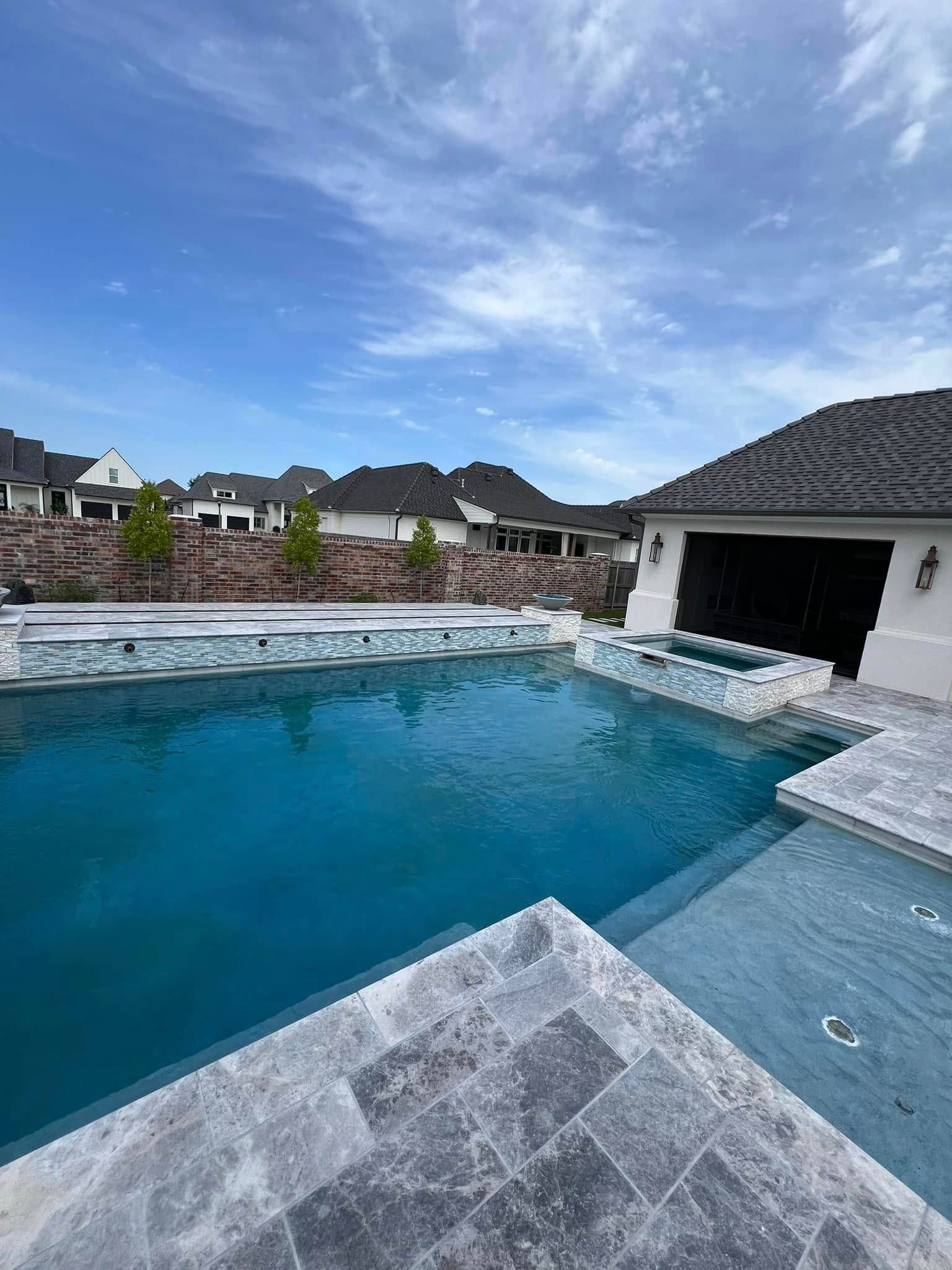 A luxury swimming pool with a hot tub and stone decking, featuring a backdrop of residential homes under a cloudy sky.