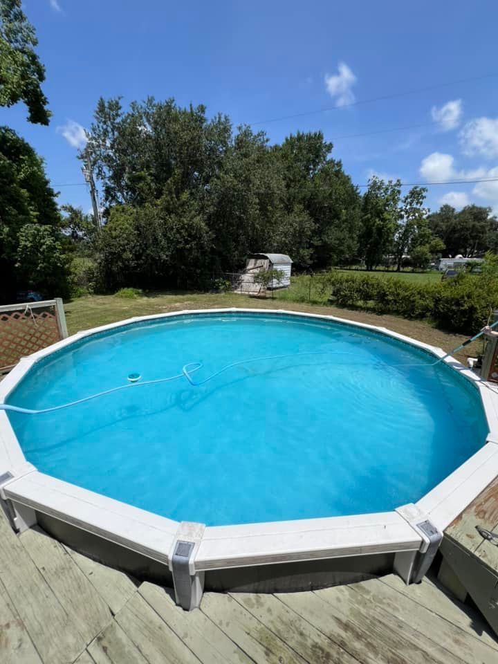 An above-ground circular swimming pool with blue water sits on a wooden deck in a grassy backyard under a clear sky.