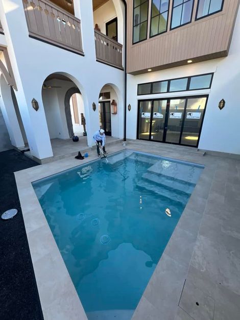 A small, rectangular swimming pool sits in a bright courtyard next to a white house with arches and dark-framed doors.