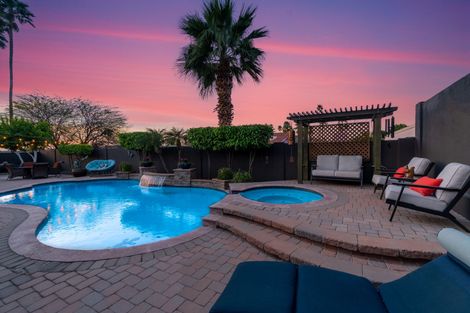 A backyard swimming pool and hot tub on a brick patio under a vibrant pink and purple sunset sky.