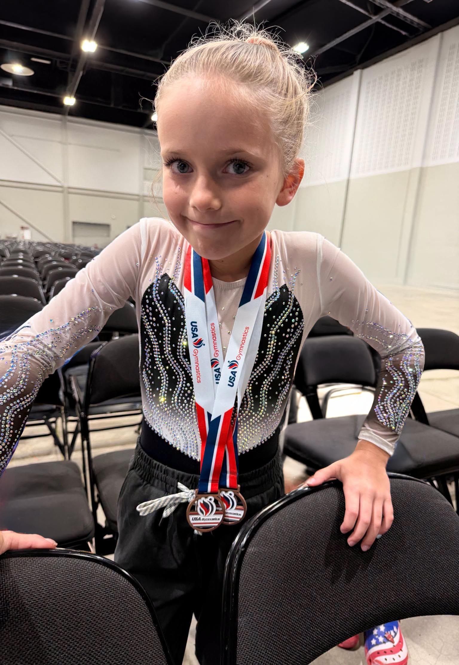 Gymnast in sparkly leotard wearing medals, seated on a chair in an indoor arena.