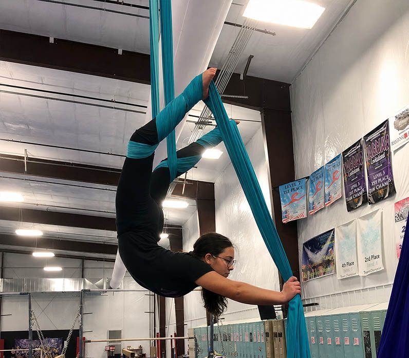Aerialist in teal silks posing upside down in an indoor gym with mats and posters nearby