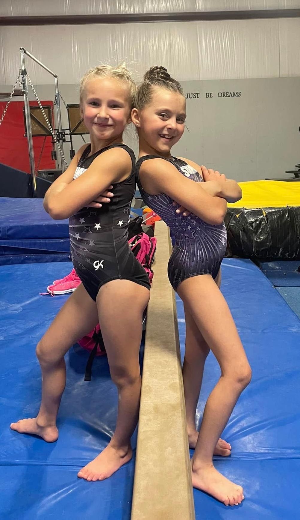 Two gymnasts posing back-to-back on a balance beam in a gym, smiling in sparkly leotards.