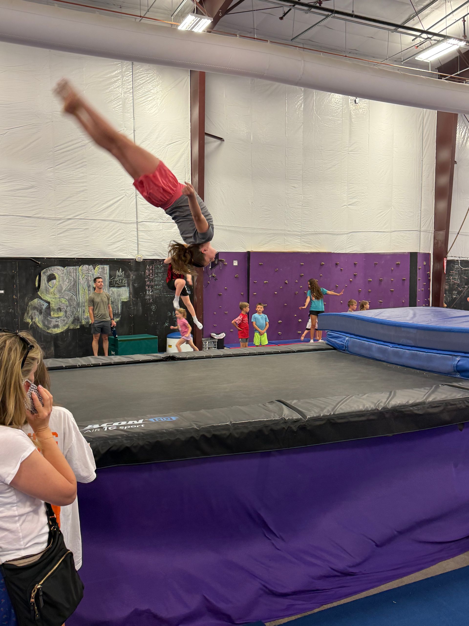 Gymnast doing a handstand on a black mat in a gym, with a purple wall and blue crash mat behind.
