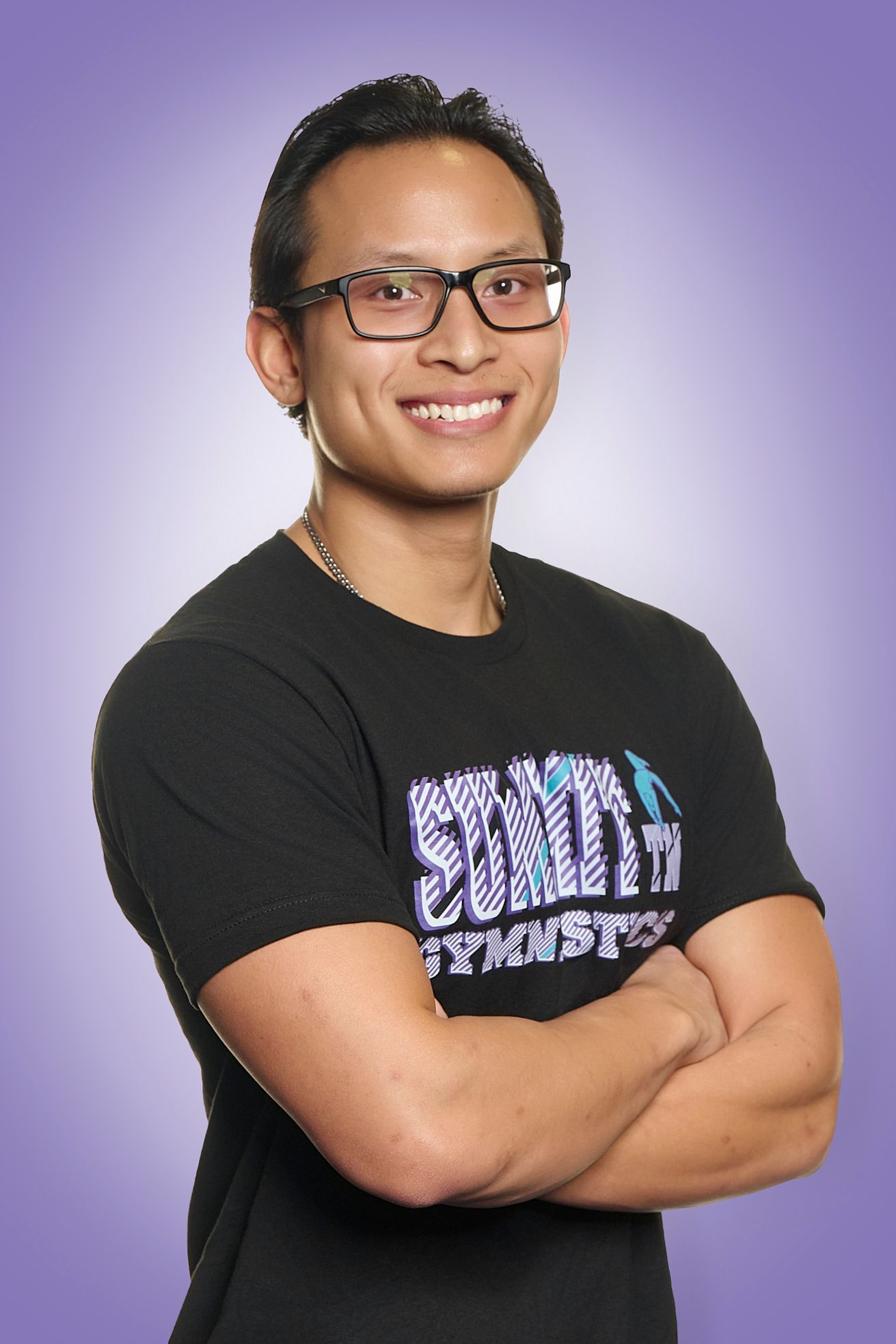 Person smiling with arms crossed, wearing glasses and a black T-shirt against a purple studio background.
