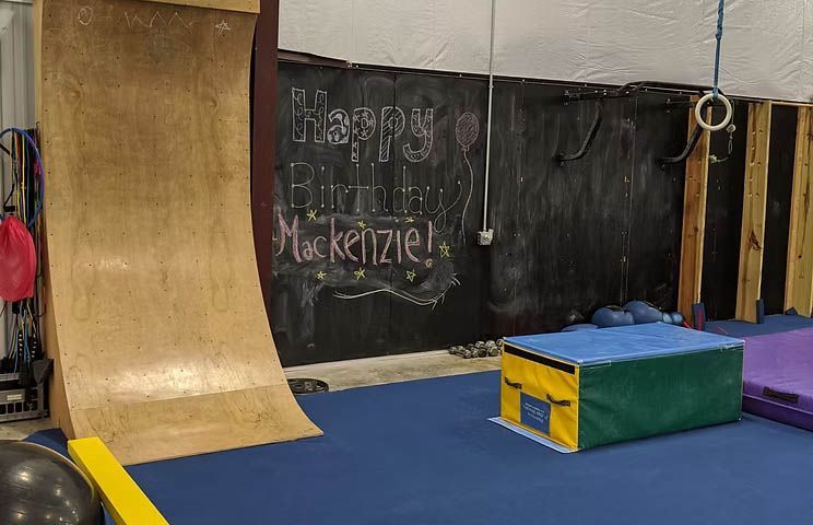 Indoor gym with a curved wooden ramp, blue floor mats, and a green padded box in front of a chalkboard wall.