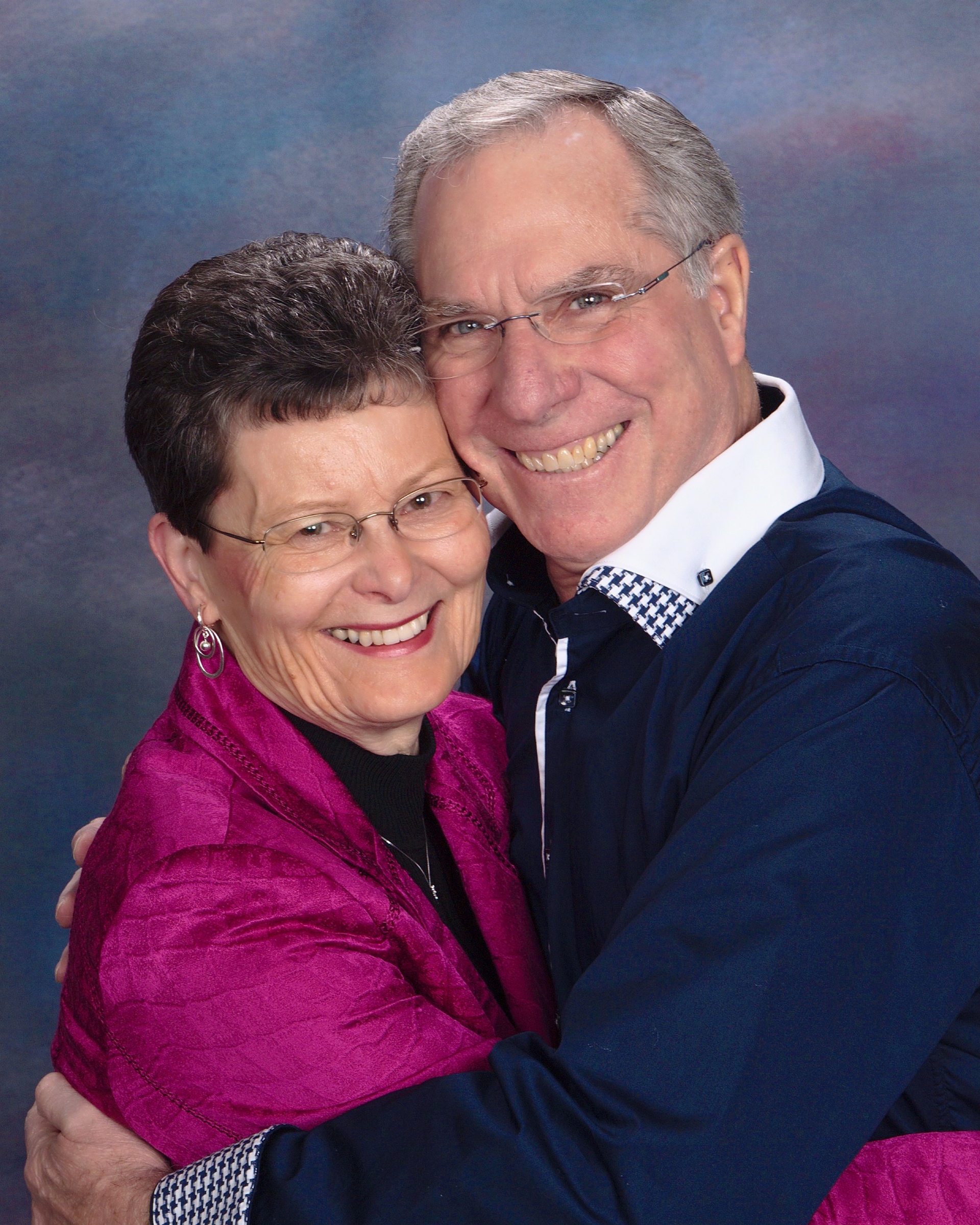 Smiling couple embraces, man wearing glasses and blue sweater, woman in pink shawl, against a blurred background.