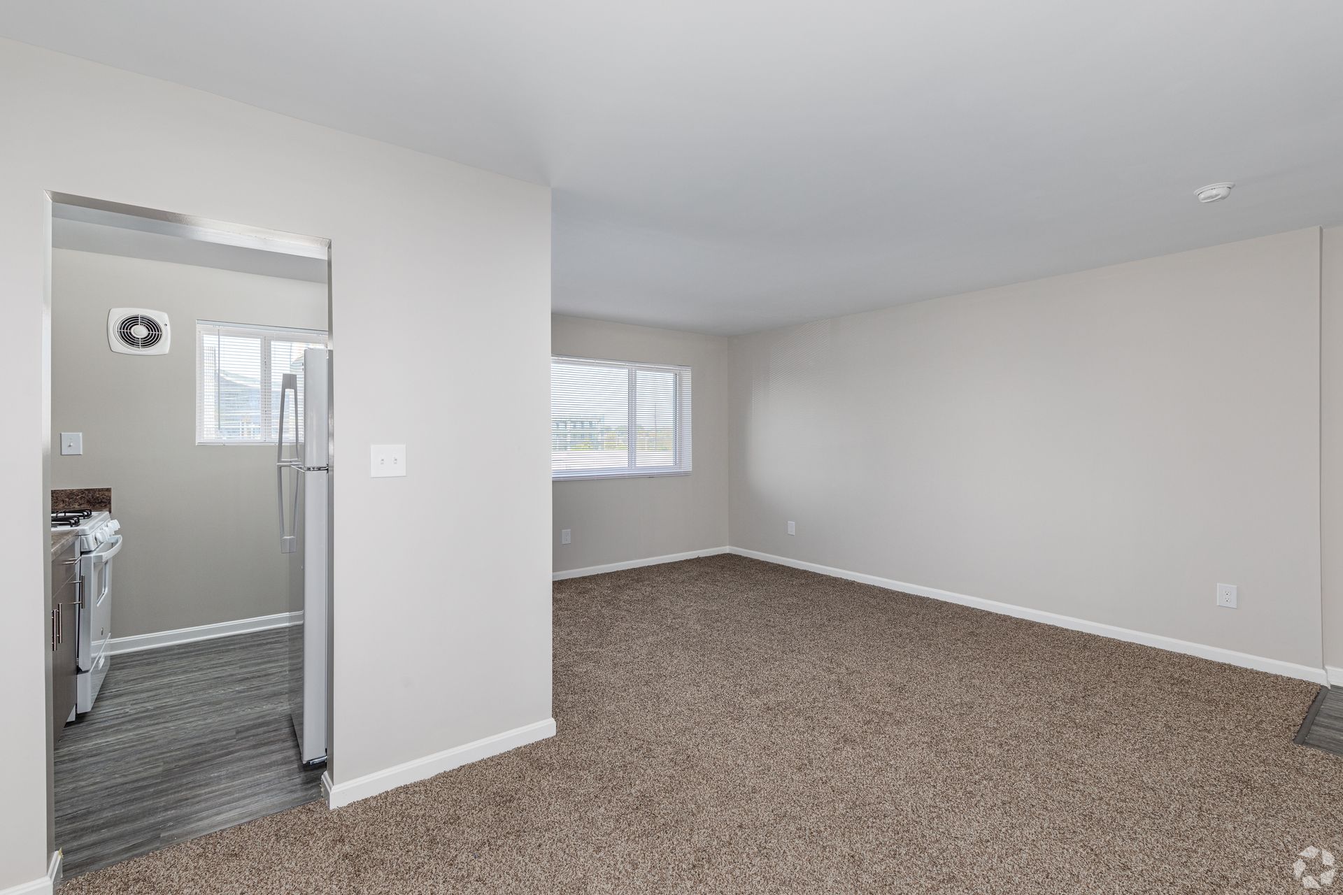 An empty living room with a carpeted floor and a refrigerator.