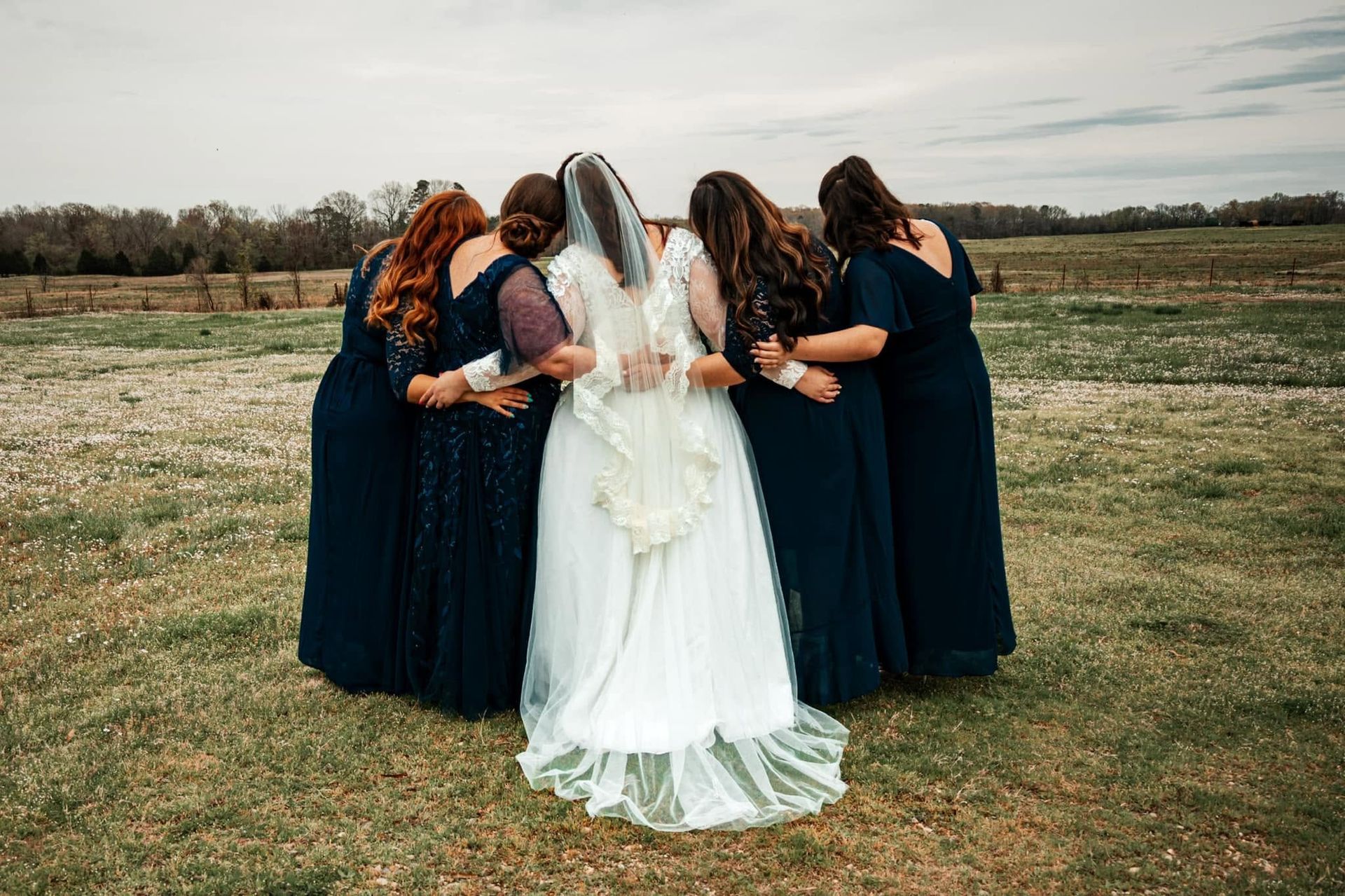 Wedding party in dark dresses surrounding a bride in a white gown outdoors on a field.