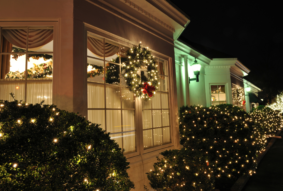 Exterior of a building lit with Christmas lights; wreath on window, bushes lined with lights.