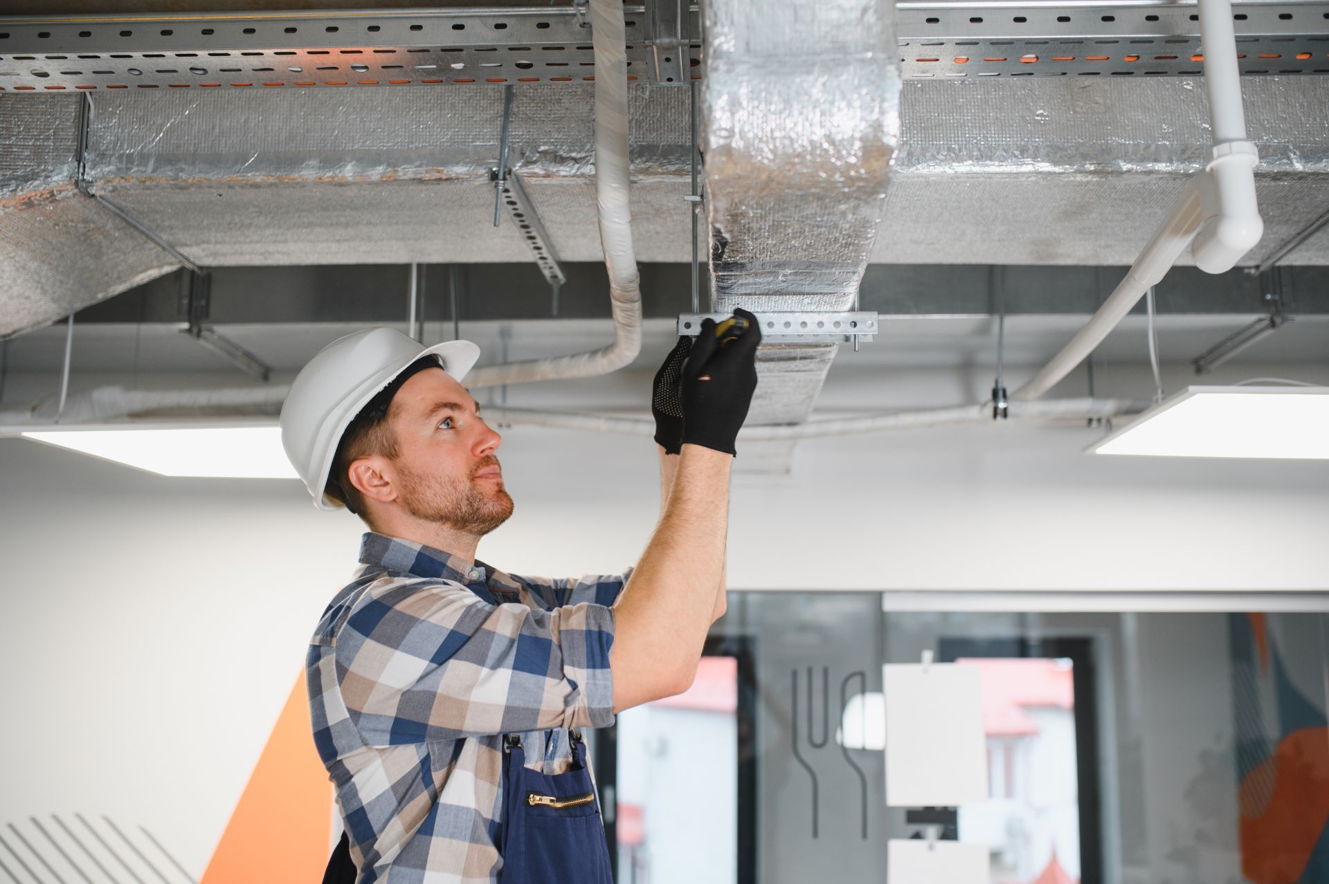 Technician in hard hat inspecting ceiling ductwork with a flashlight in a commercial interior