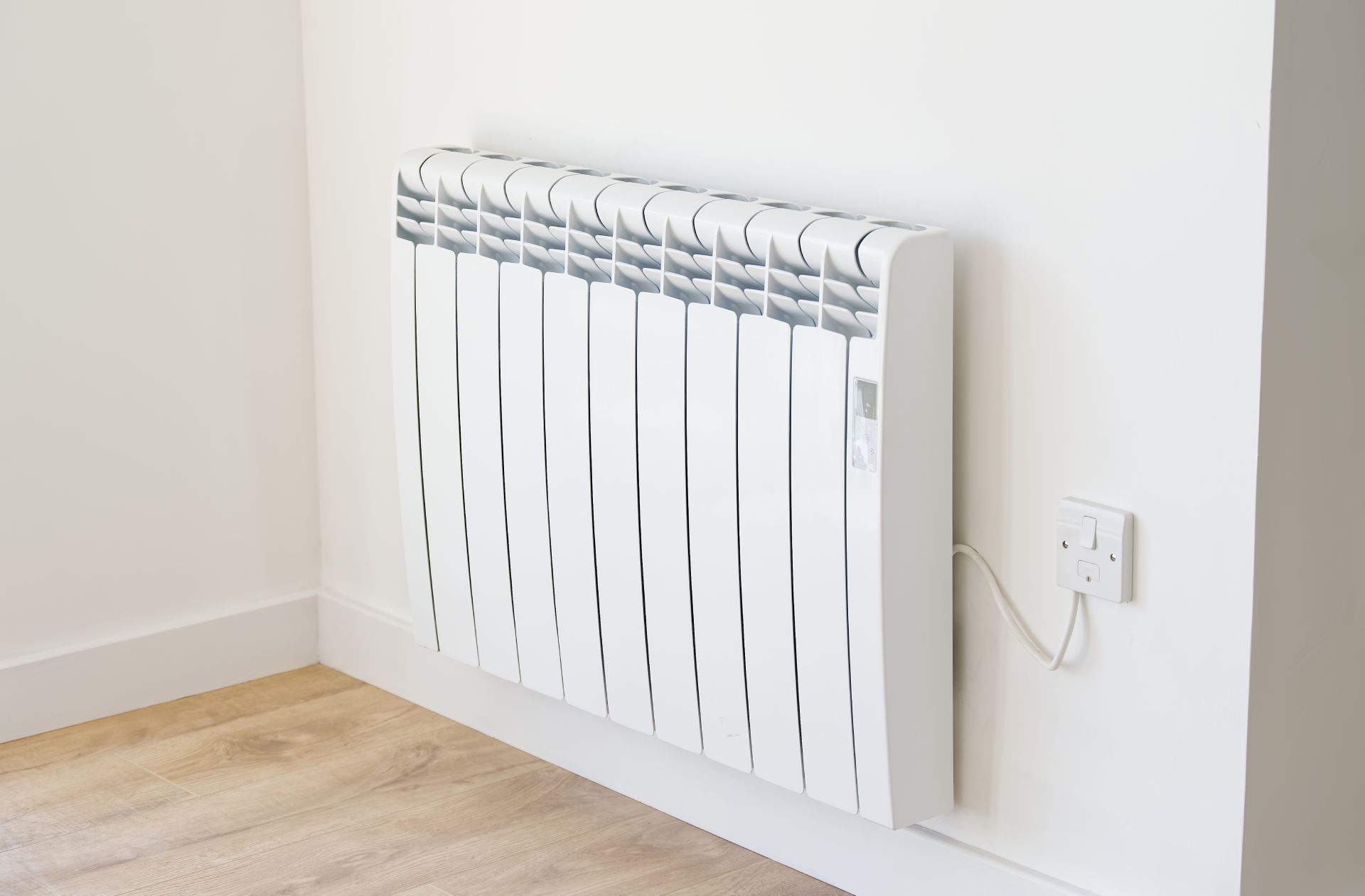 White wall-mounted electric radiator with cord on a light wood floor against a plain wall