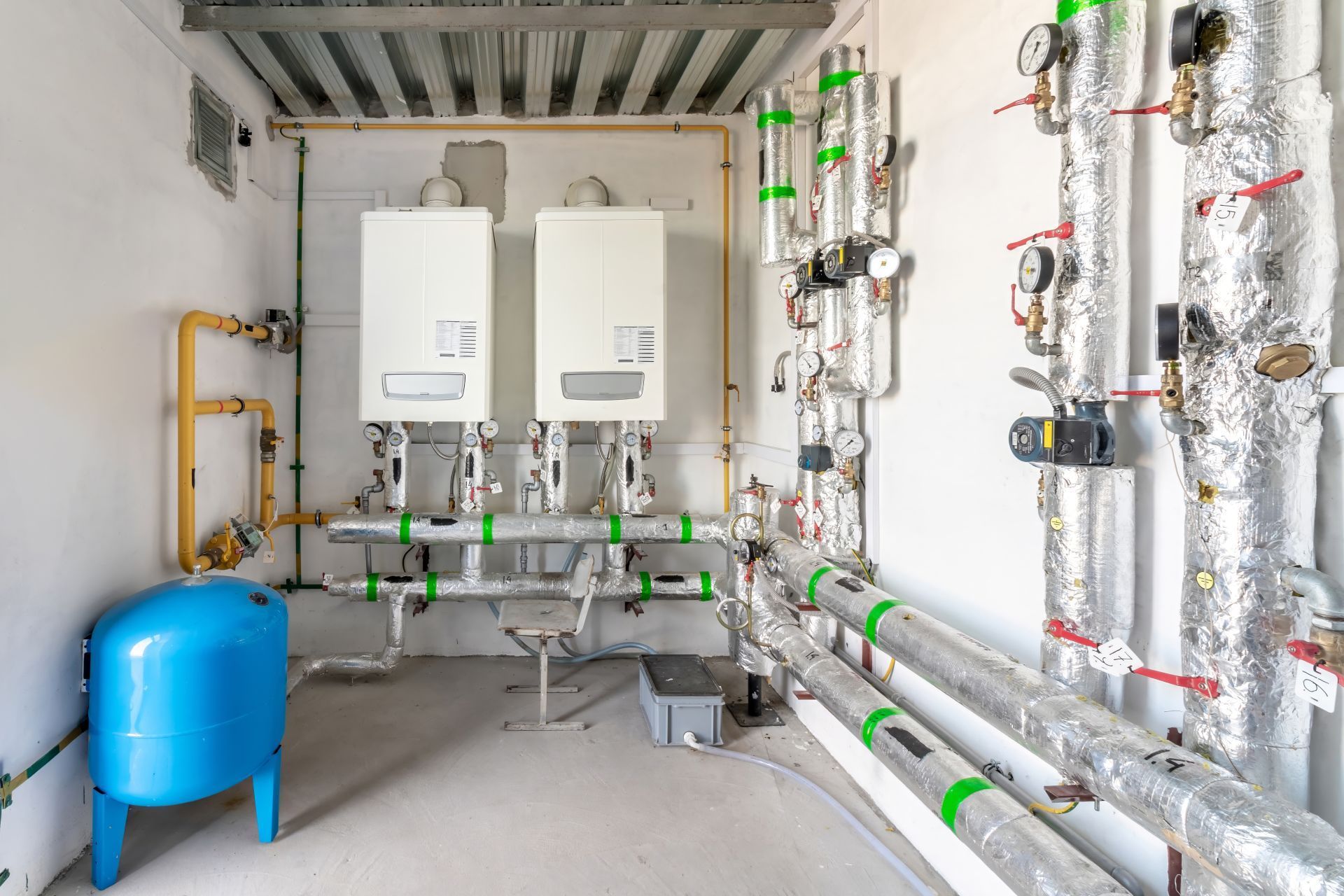 Utility room with two wall-mounted boilers, exposed pipes, and a blue expansion tank