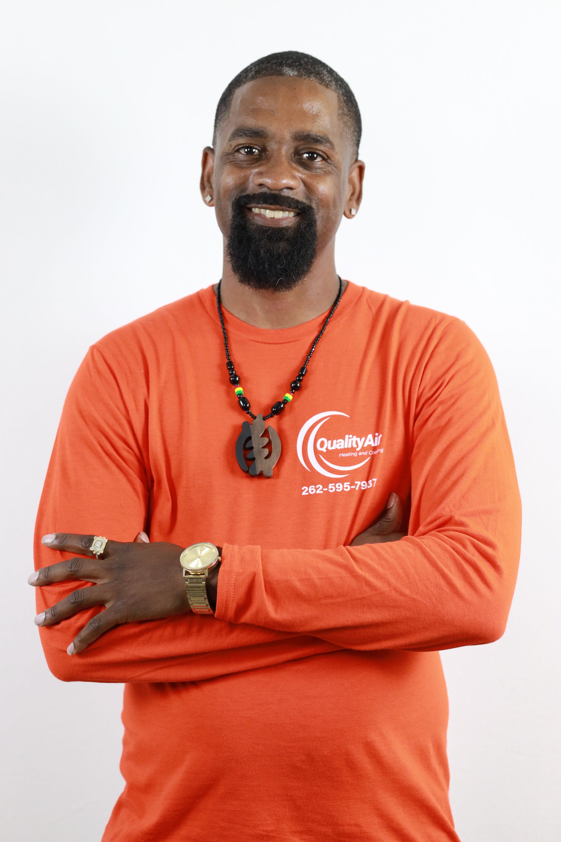 Man with crossed arms, wearing an orange long-sleeved shirt with a logo, necklace, and rings; smiling against a white backdrop.