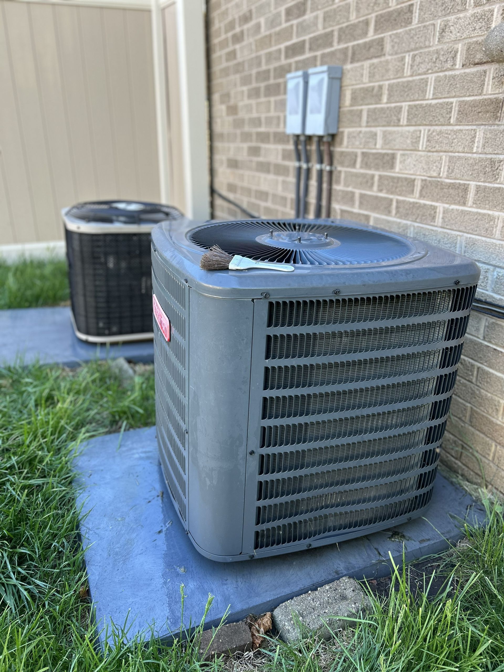 Two air conditioning units on concrete pads next to a brick wall and grass.