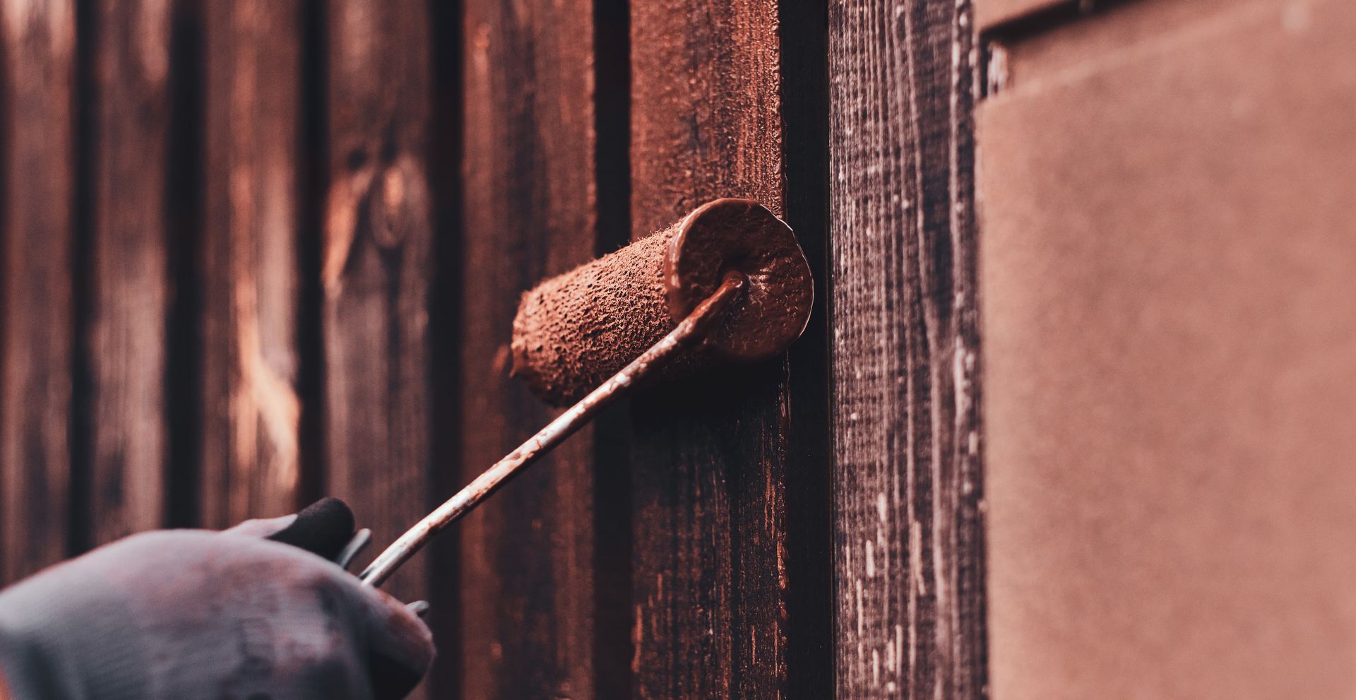 A gloved hand uses a paint roller to apply dark stain to a wooden fence.