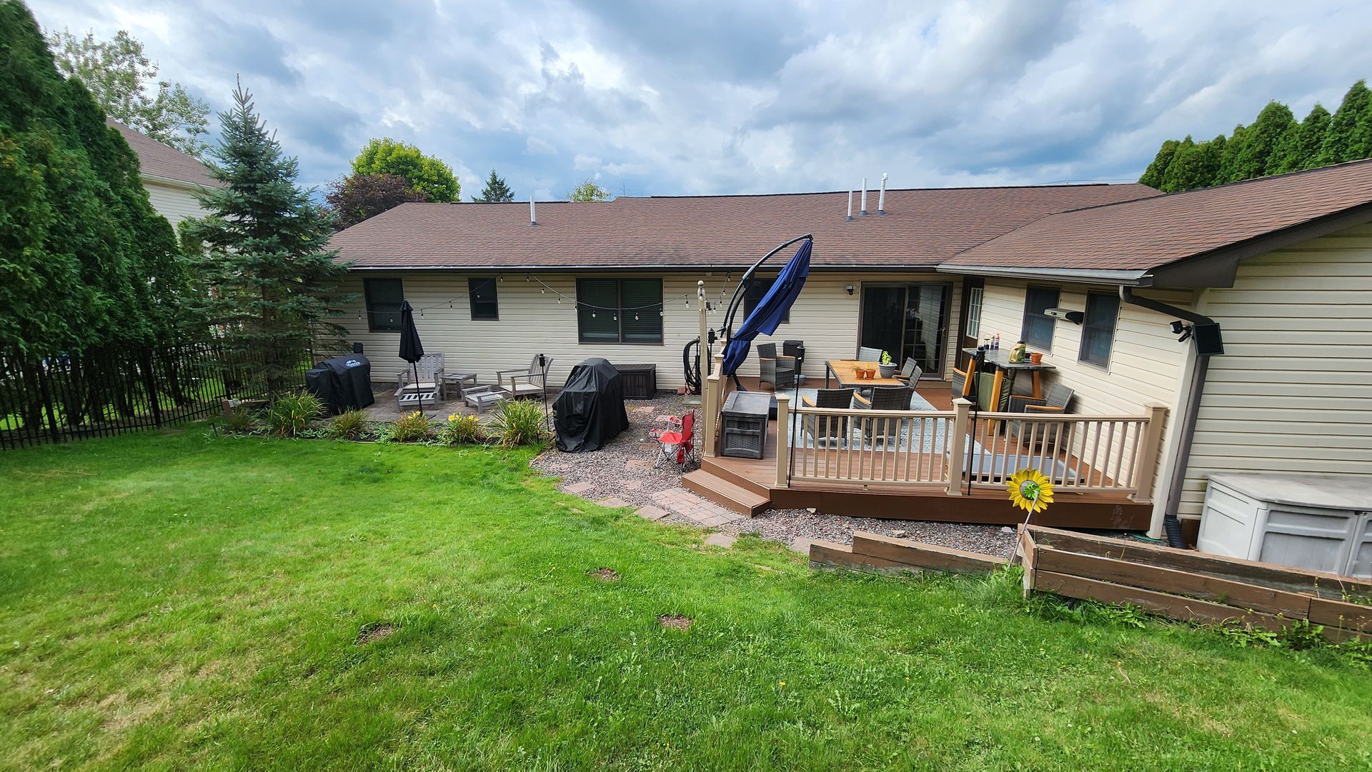 Backyard with a wooden deck, grill, and brown-roofed house. Green lawn and cloudy sky.