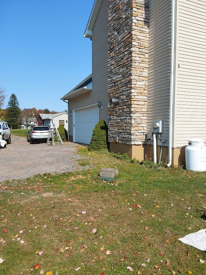 Side view of a beige house with a garage, a stone chimney, and a propane tank, set on a grassy lawn.