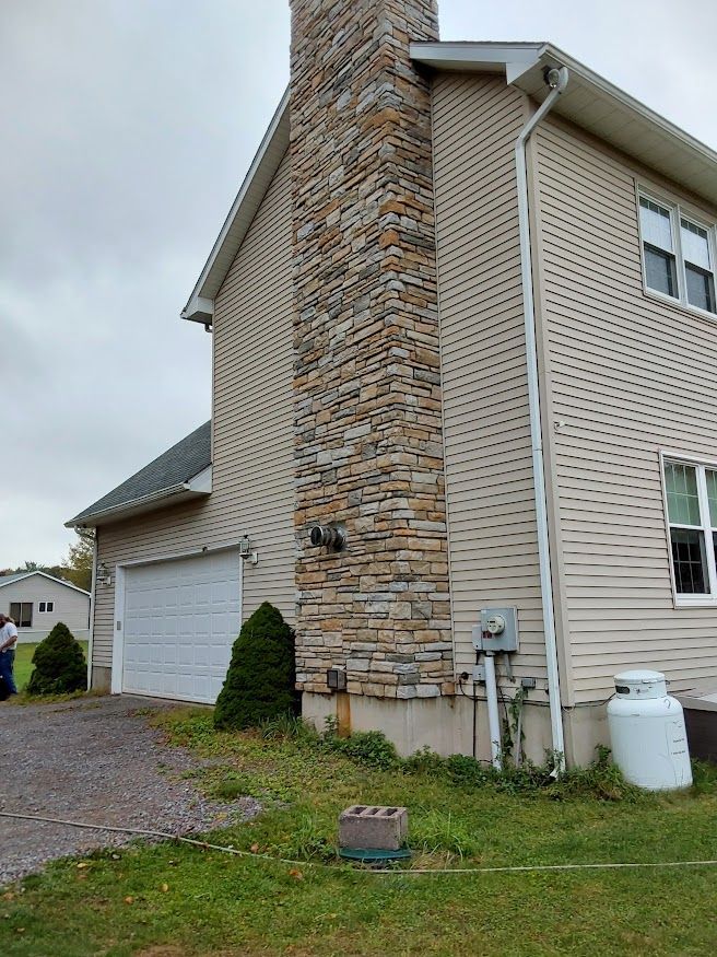 Side view of a house with stone chimney and siding. A garage door and propane tank are also visible.