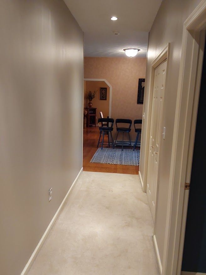 Narrow hallway with beige walls, carpet, and doors leading to a room with chairs.
