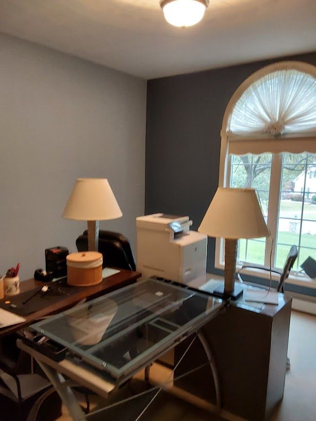 A home office with a desk, lamps, a printer, and a window with a beige curtain. Blue walls.