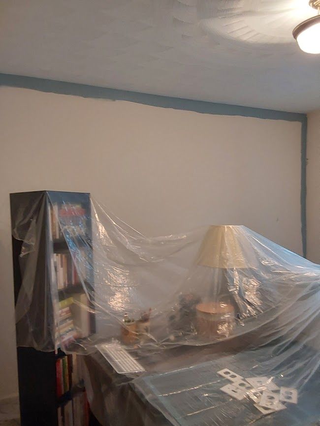 A room with a blue-trimmed white wall, partially covered in plastic. A bookshelf and desk are also covered.