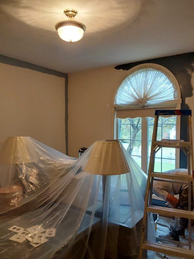 Room with covered furniture, a ladder, and a person working near a window with a half-circle shade.