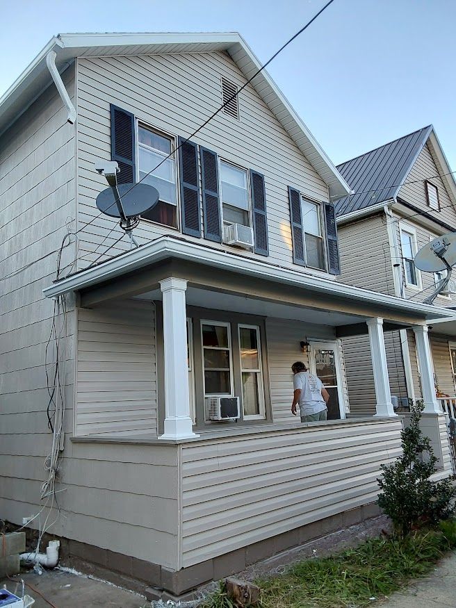 Two-story house with porch; light beige siding, dark shutters, satellite dishes. Person in doorway.