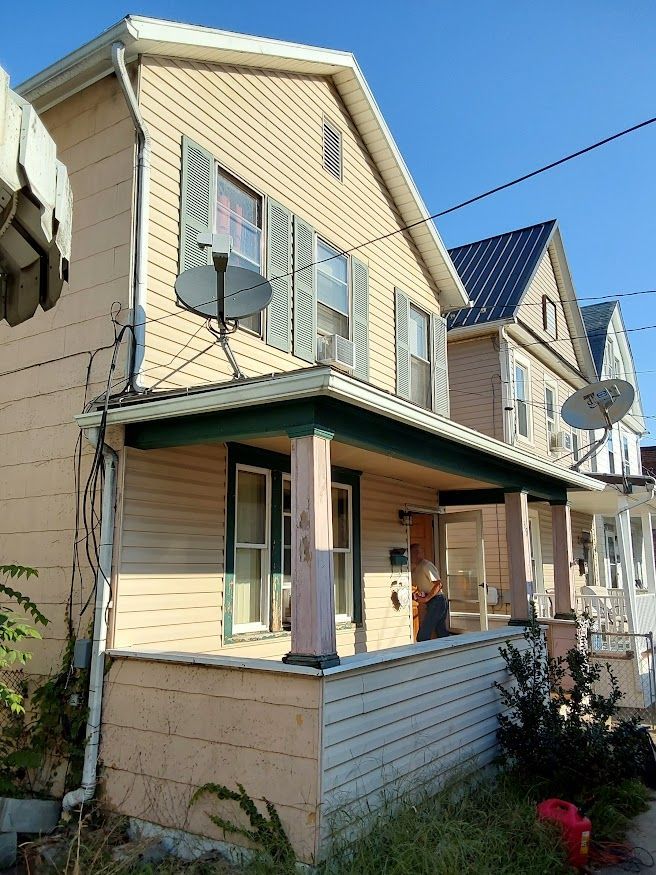 Two-story house with a porch. Beige siding, green trim, two satellite dishes, blue sky.