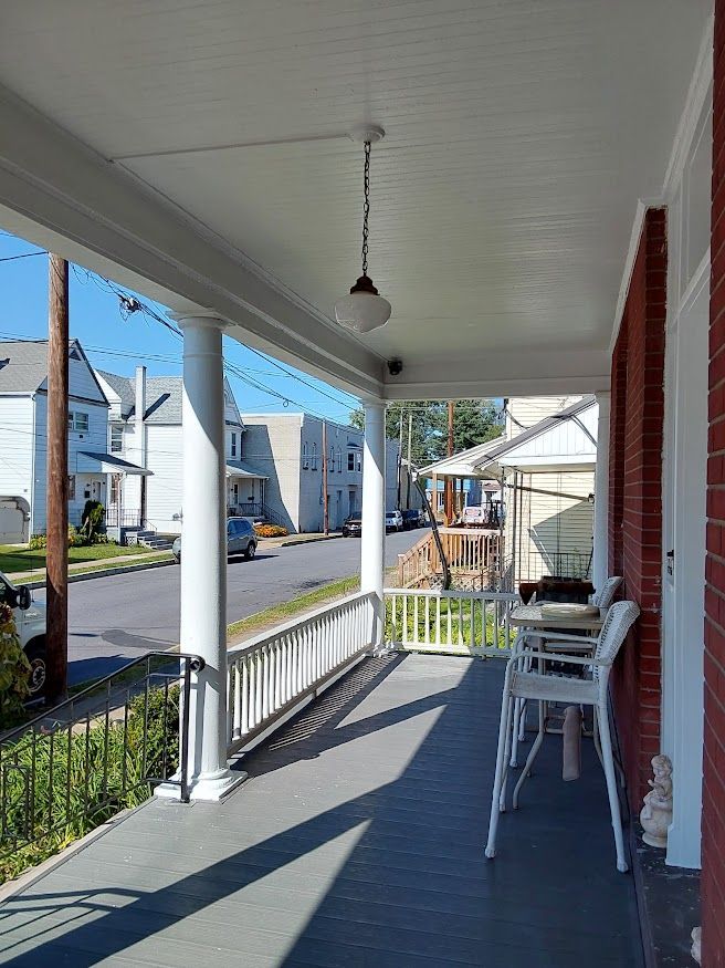 A porch with white columns, railing, and ceiling. Two white chairs sit near a red brick wall, overlooking a street.