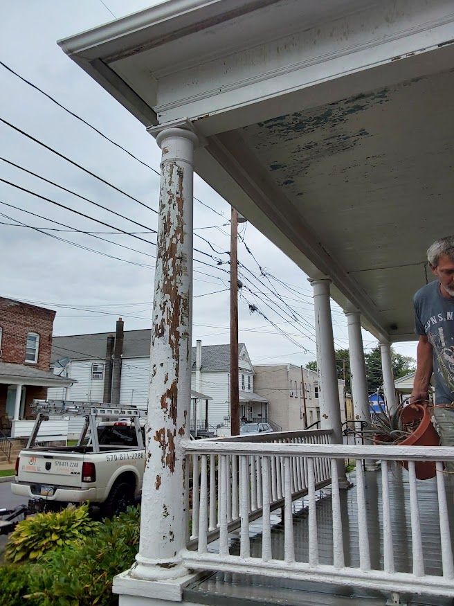 Porch with peeling paint on a column, man holding object, truck parked on street, overcast sky.