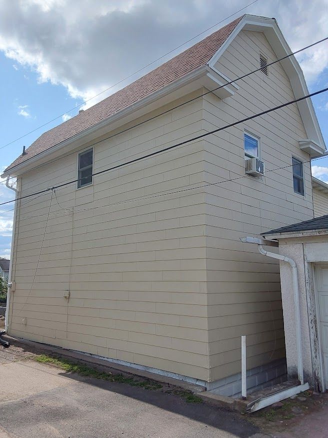 Side of a two-story beige house with a brown roof and small windows, set on a paved area under a cloudy sky.