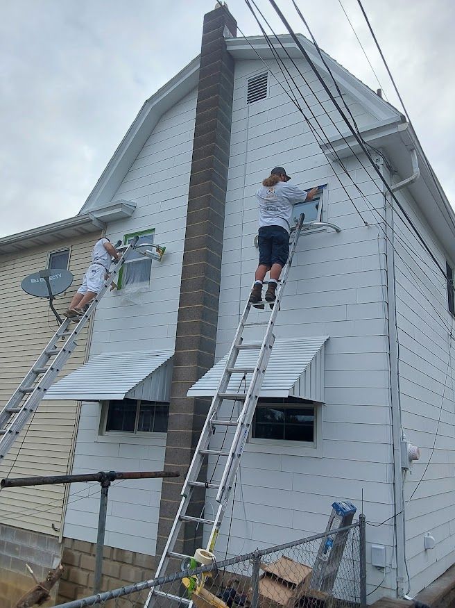 Two people on ladders installing something on the side of a white house with a chimney.