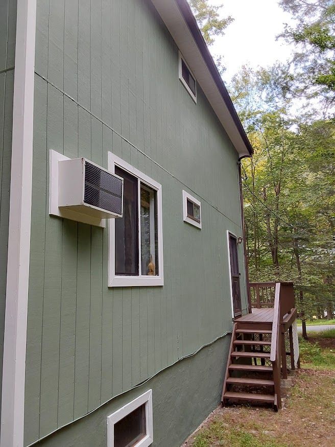 Green house with white trim, air conditioner in window, wooden steps leading up to a door.