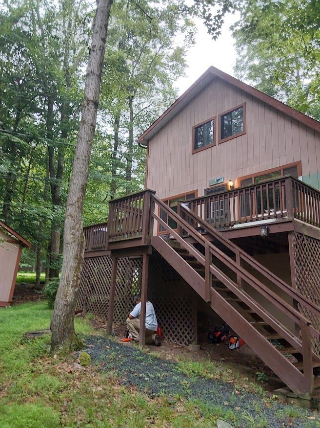 Brown house with a wooden deck. A person crouches beneath the deck. Trees surround the house.