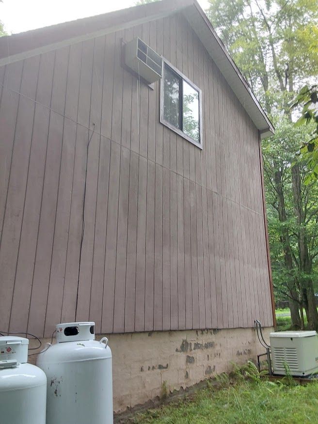 Side of a house with vertical brown siding, a window, and propane tanks. A generator sits in the grass.