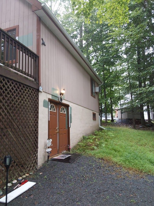 Exterior of a building with brown doors, a deck, and an unpaved area. Trees line the right.