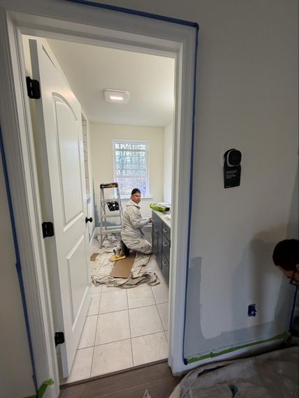 Hallway view into a room with a worker in white coveralls, tools, and unfinished flooring.