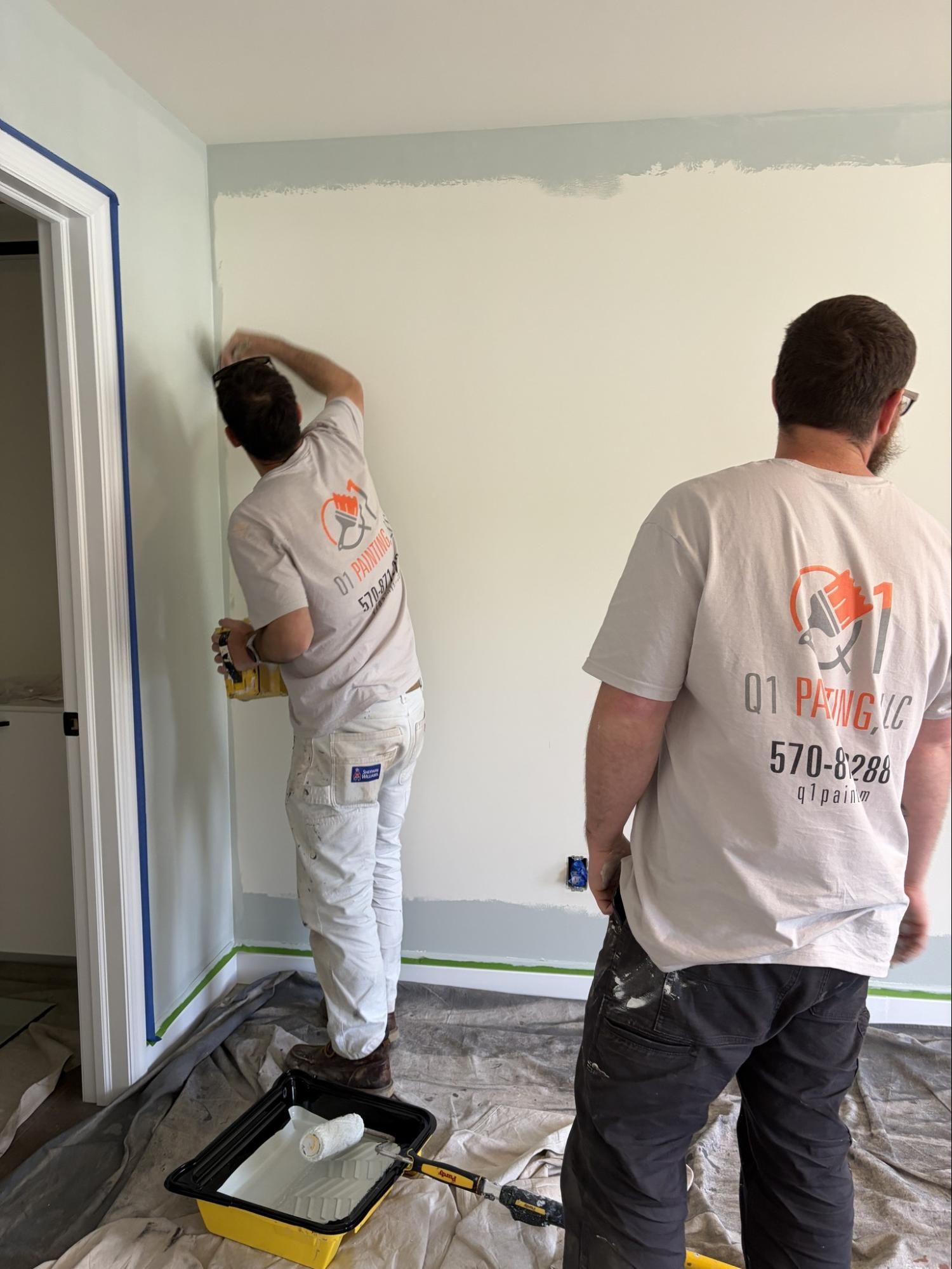 Two workers painting a white room, one rolling paint near the wall and one standing by the doorway.