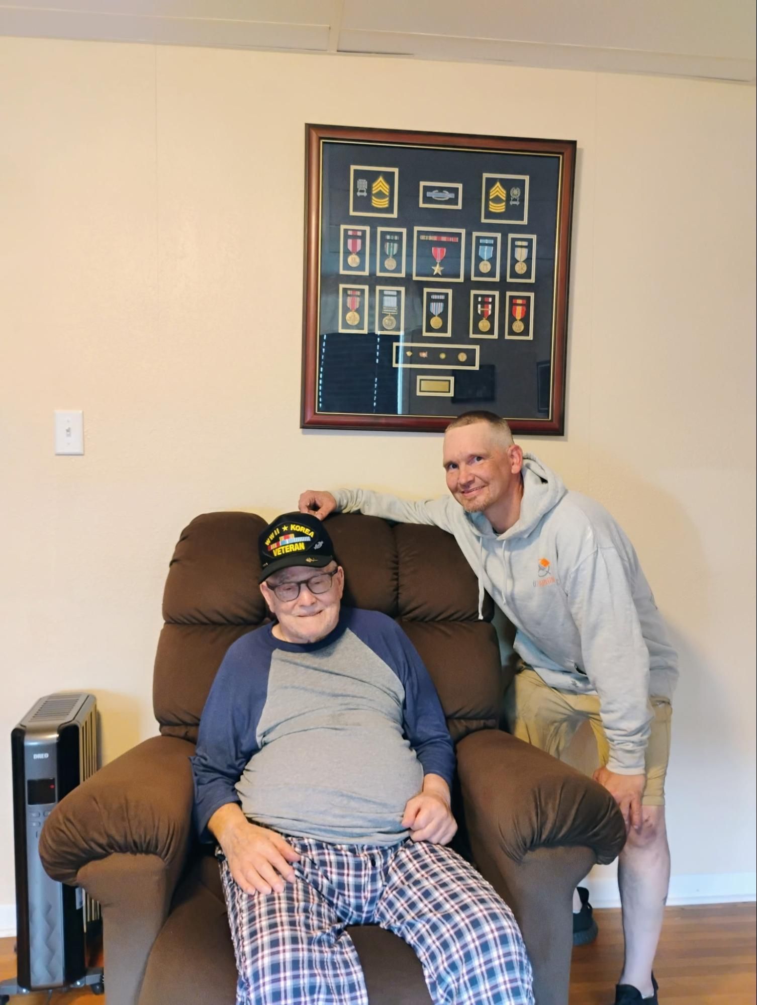 Two people posing beside a seated man in a recliner, with a framed display on the wall behind them.
