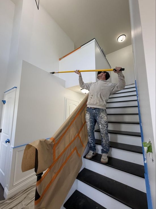 Worker painting stairwell with taped-off walls and plastic sheeting, standing on stairs with a long roller pole